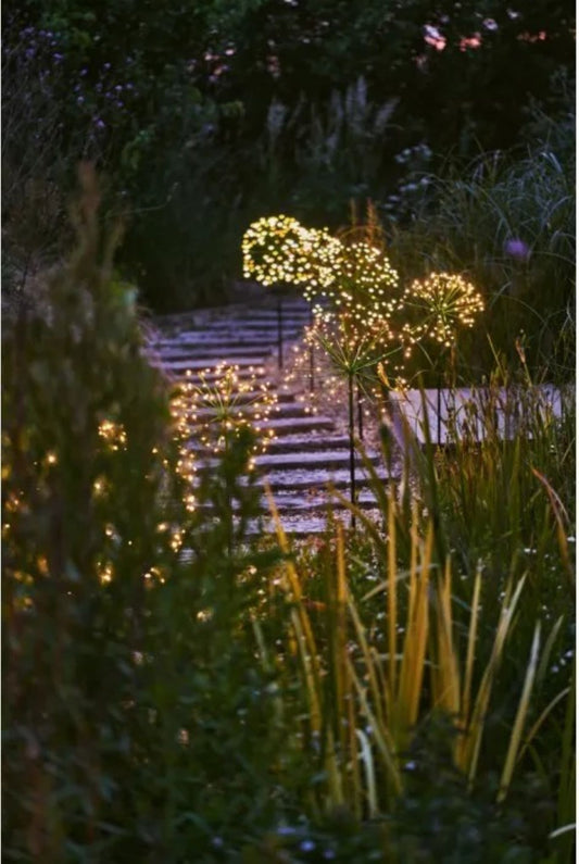 stor solcellelanterne på spyd plassert i blomsterbed med varmt lys