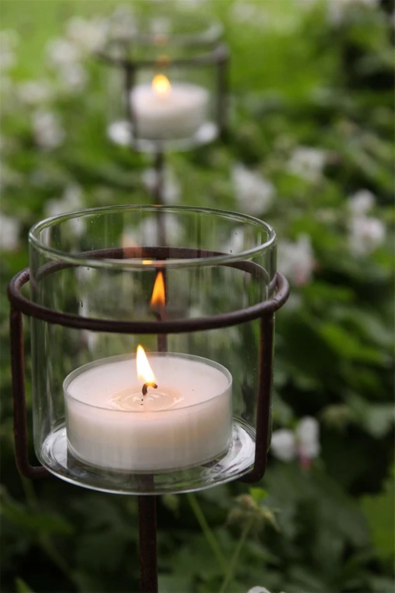 Høy telysholder med glass plassert i blomsterbed i hagen